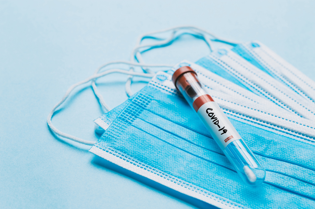 One single glass test tube containing a Corona virus test swab sitting on top of some blue surgical masks